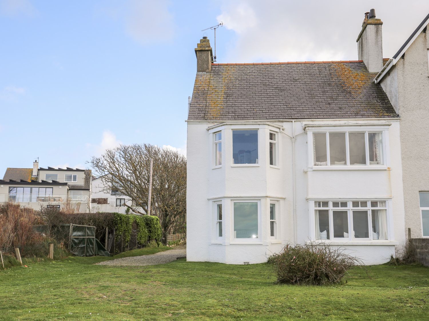 The exterior of a white two-story house with multiple windows and a sloped roof with chimneys surrounded by grass and shrubs at Windygates in Rhosneigr