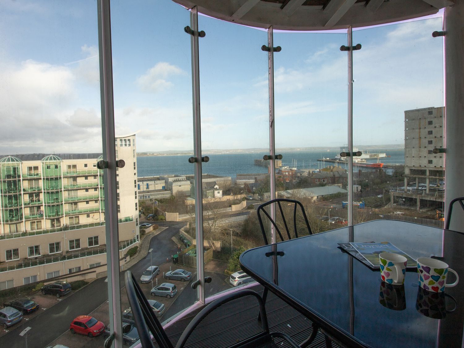 A balcony with glass table and chairs overlooking buildings and a body of water at Panoramic View in Castletown Portland