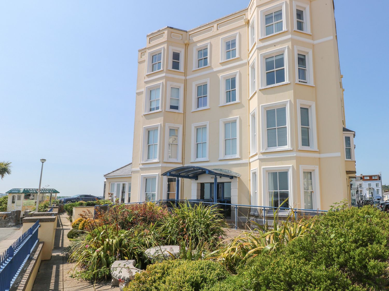 A beige multi-story building with white-trimmed windows surrounded by plants and a blue fence at 6 South Beach Court in Tenby