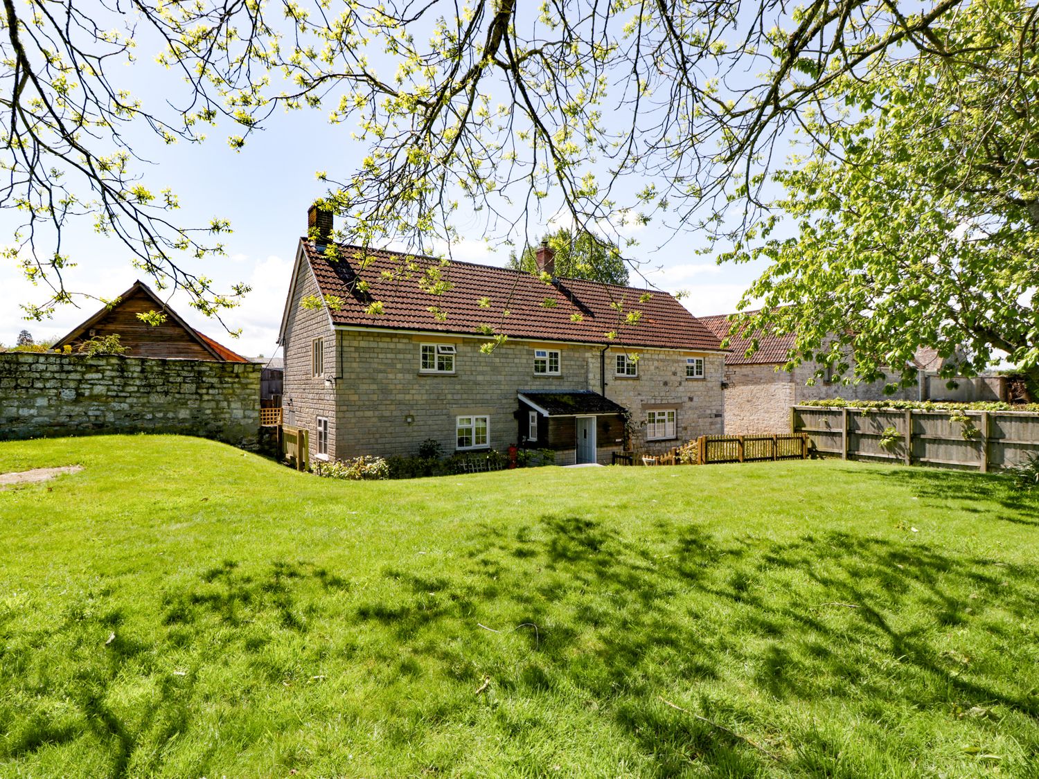 A stone house with a tiled roof and multiple windows surrounded by a lawn and trees at The Stable House in Sherborne