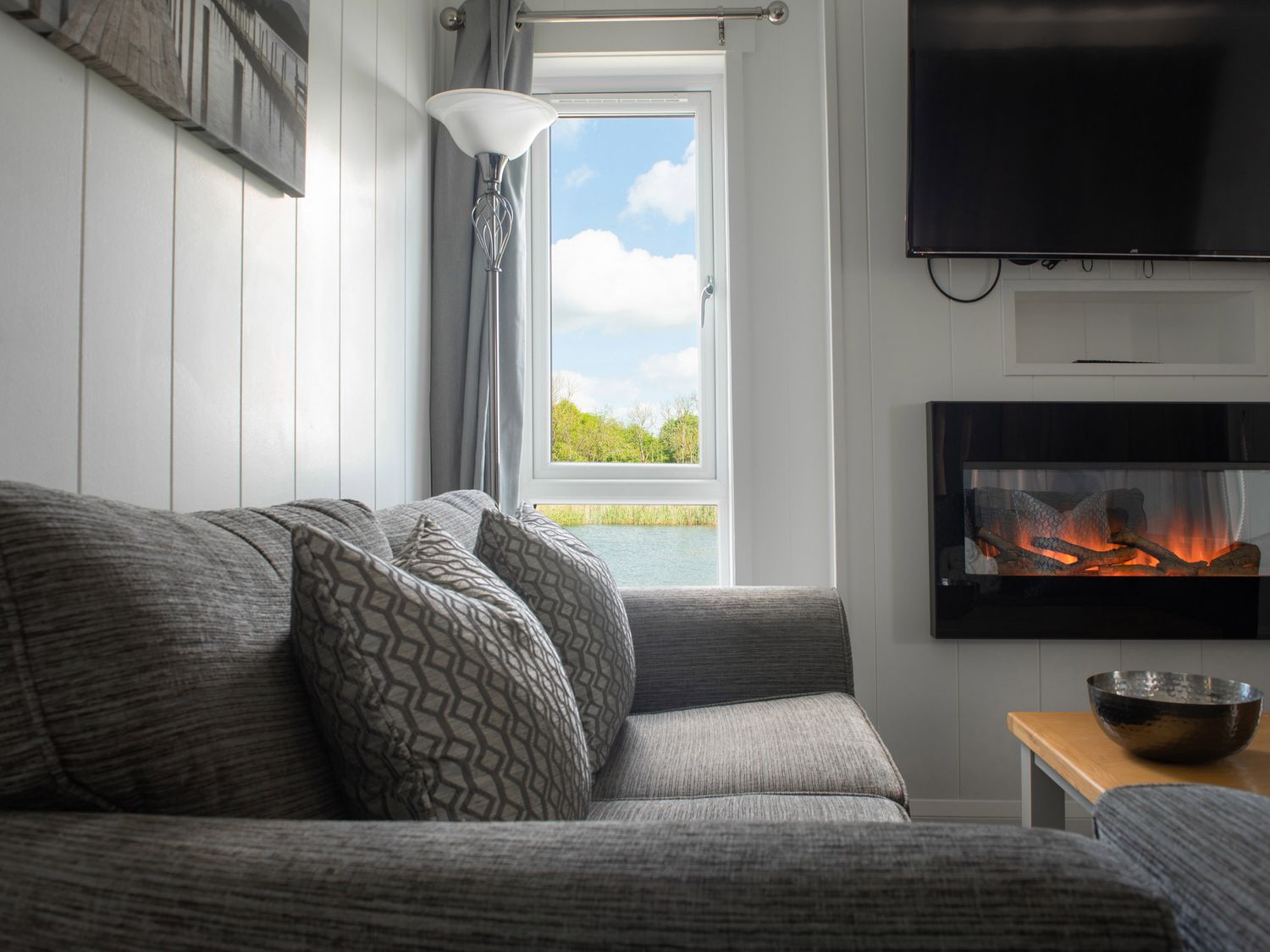 A living room with a gray sofa and pillows a window with a view and a mounted electric fireplace at Lodge 7 - The Earn in Errol