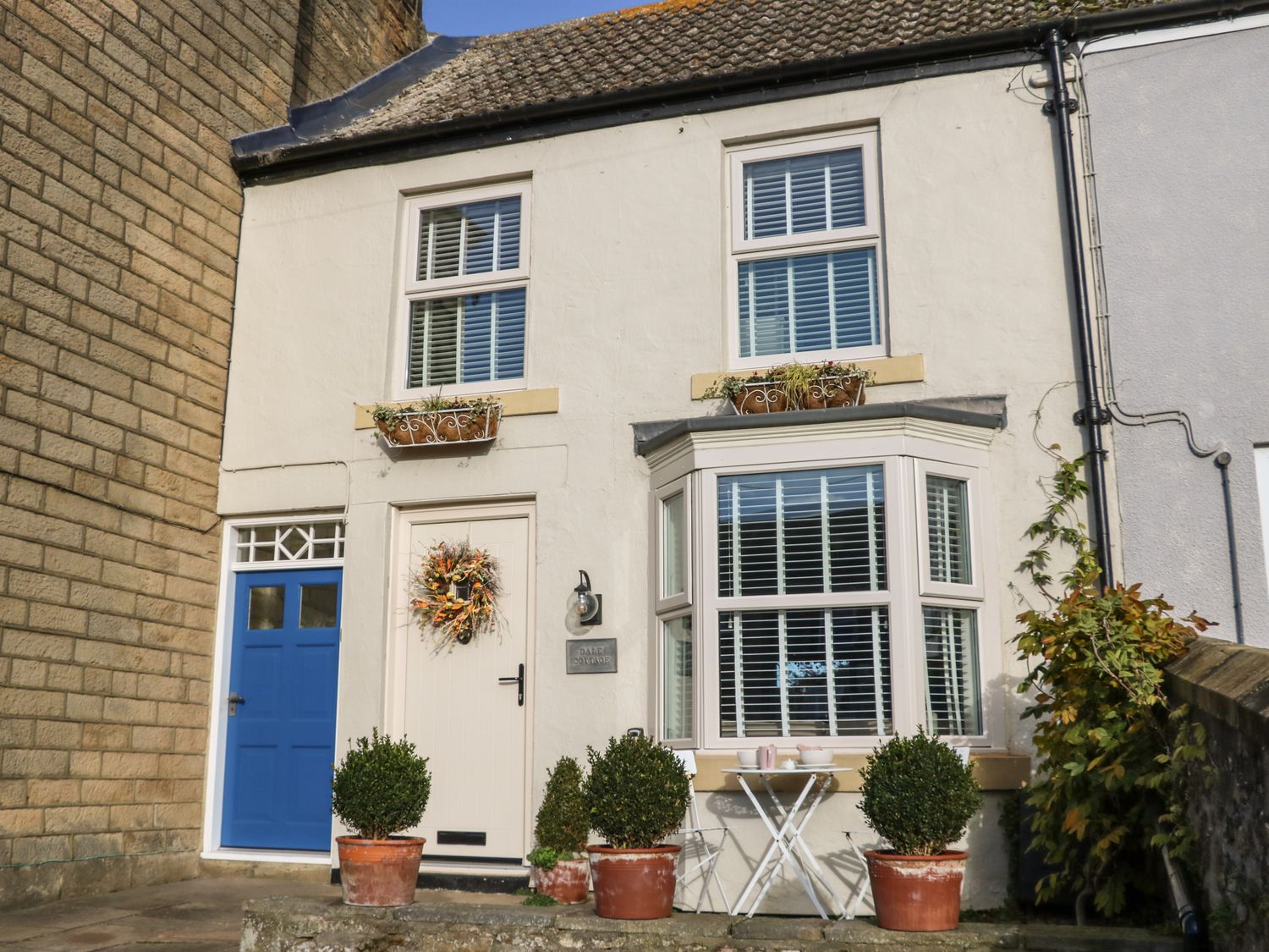 White two-story house with blue door white door with wreath bay window and potted plants on the front patio at Dale Cottage in Ingleton County Durham