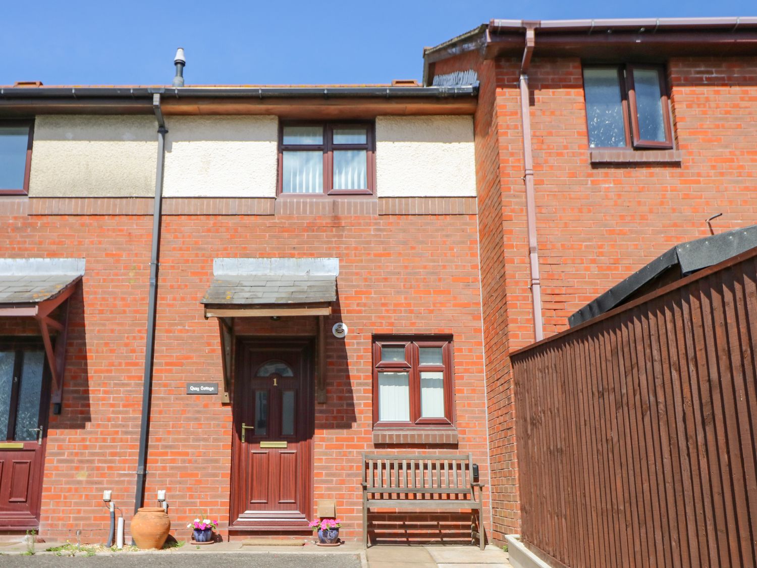 A red brick exterior of a house with a maroon front door and windows a wooden bench and flower pots outside at Poole Quay Cottage in Poole