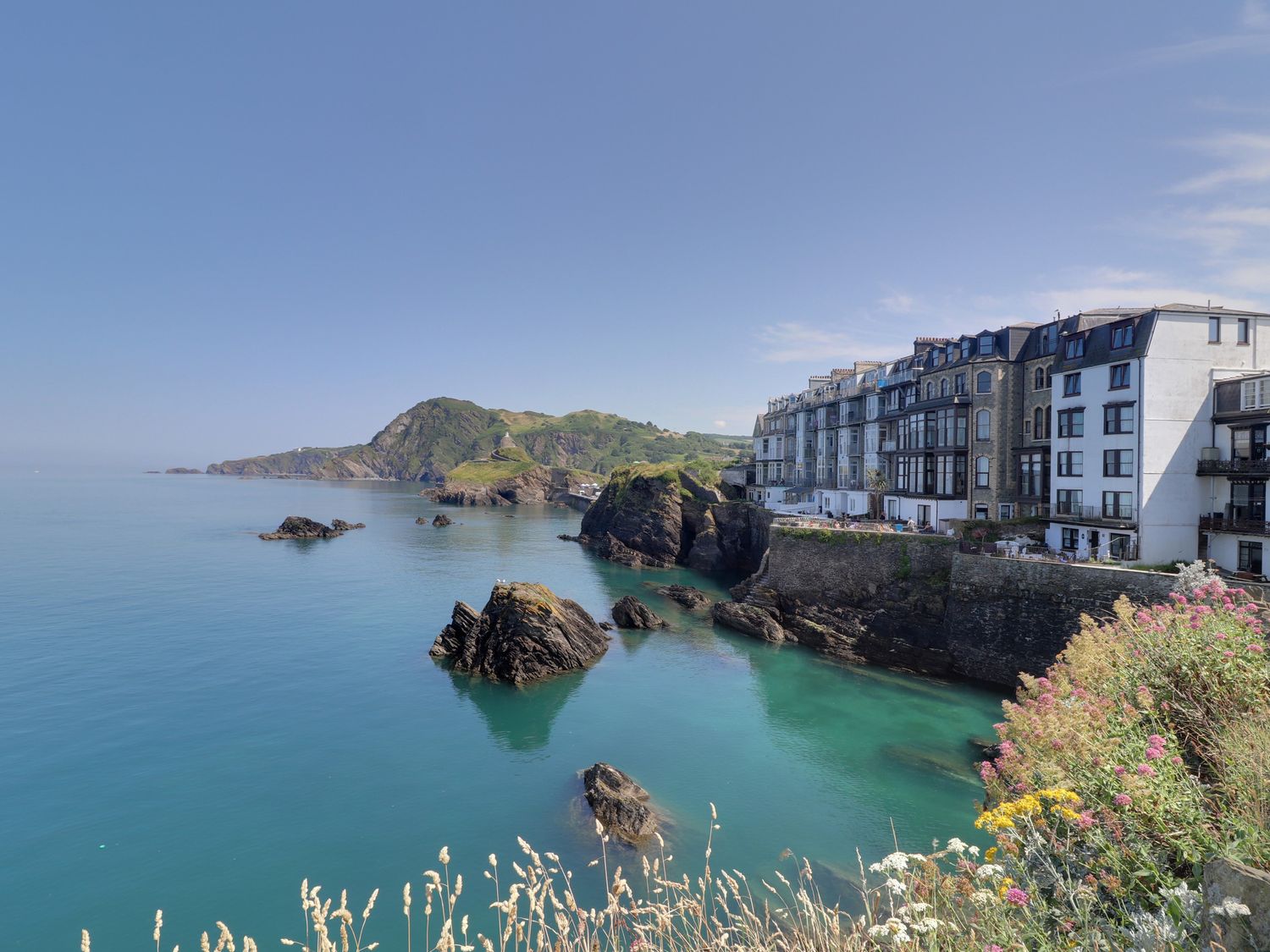 A coastal scene with buildings on a cliff overlooking rocks in the sea at 1 Cheyne Beach Apartments in Ilfracombe