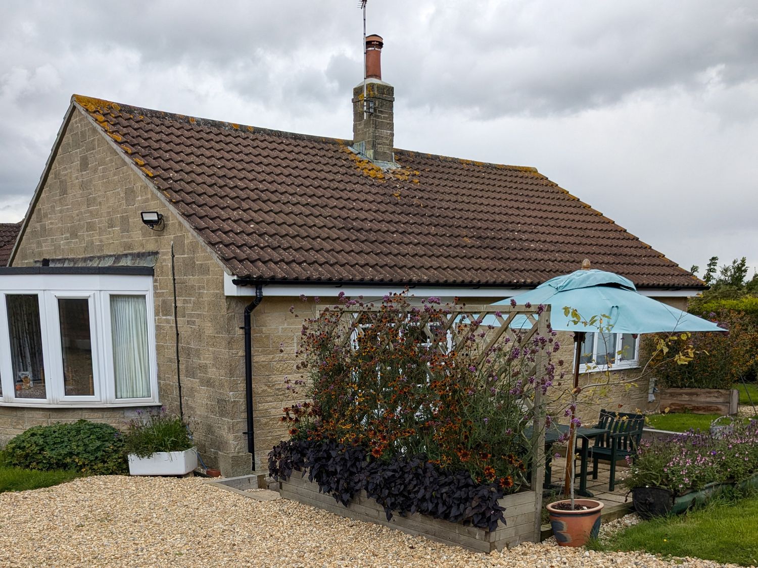 A garden with flowers and a blue umbrella next to a stone house with a tiled roof at Rhode Acres in Fifehead Neville near Hazelbury Bryan