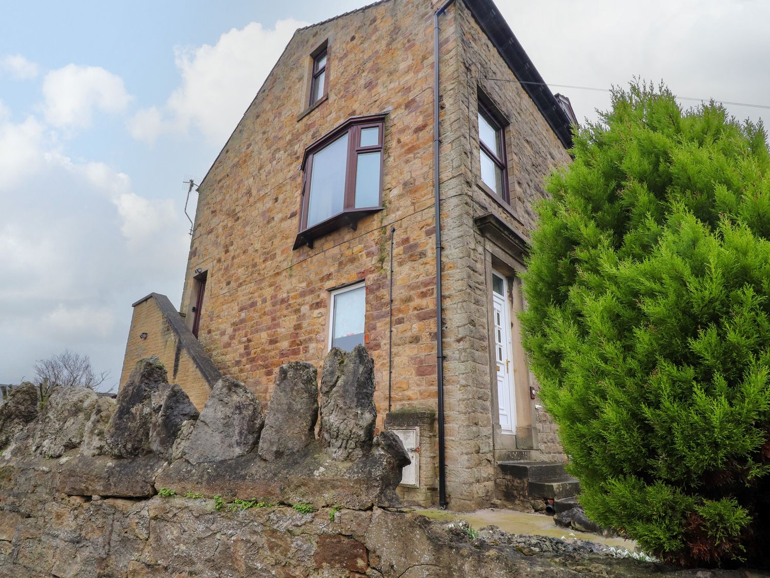 A stone house with a white door and windows next to a stone wall and a green bush at Mountain View in Hest Bank near Slyne