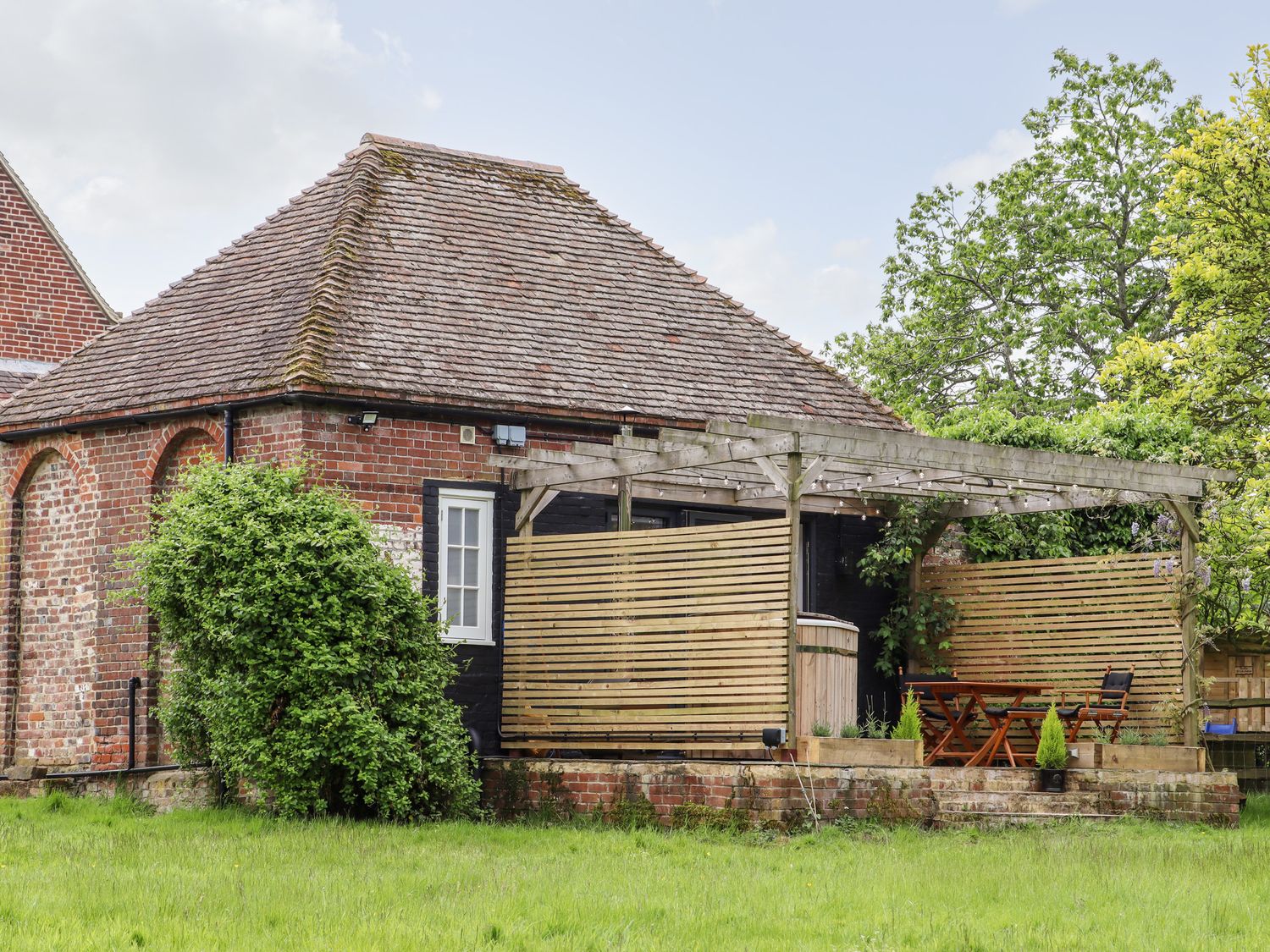 A brick building with a tiled roof a wooden pergola with outdoor furniture and plants in a grassy area at The Snug at Pickelden Farmhouse Mystole near Chilham