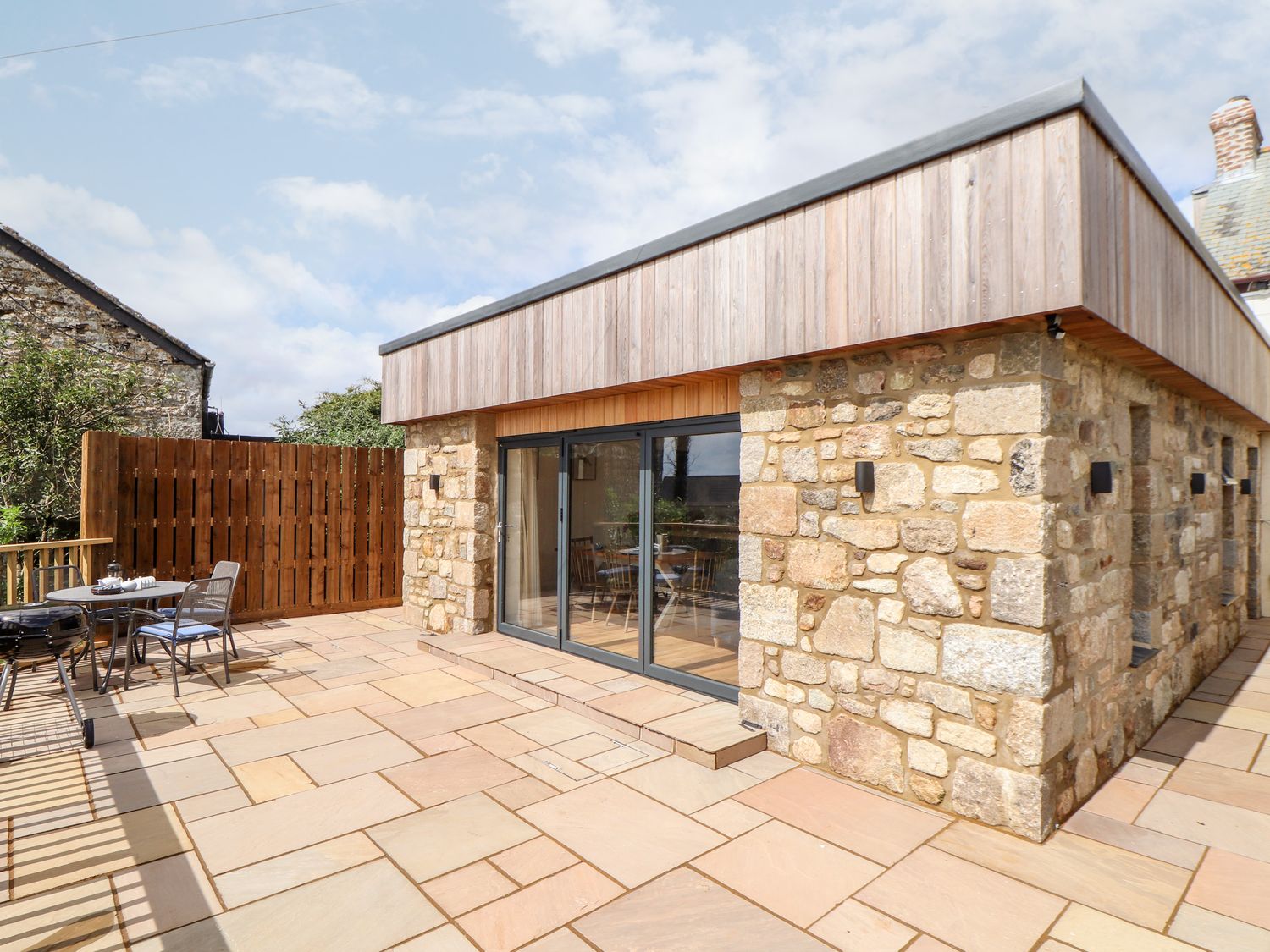 A stone building with wooden siding and patio furniture on a tiled terrace at Chi an Mor in Lower Kenneggy near Praa Sands