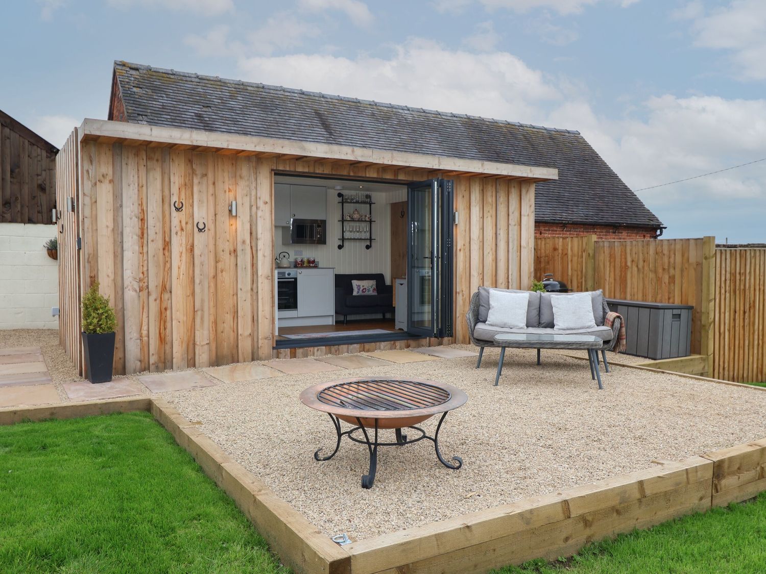 An outdoor seating area with a small fire pit and a sofa in front of a wooden cabin with open doors showing a kitchenette at Hillside Hut in Radbourne near Ashbourne
