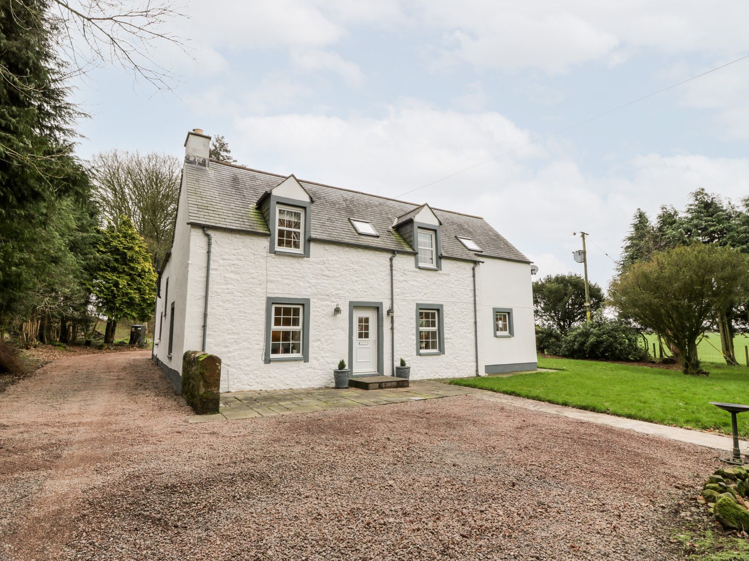A white stone house with gray trim and a gravel driveway surrounded by trees and grass at Oakbank in Carrutherstown near Annan