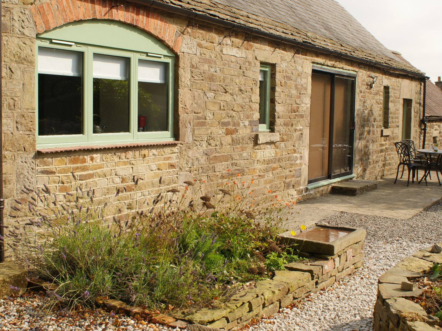 A stone exterior wall with green framed windows and a sliding glass door next to a garden bed and outdoor seating area at Temperance Croft Wadshelf near Chesterfield