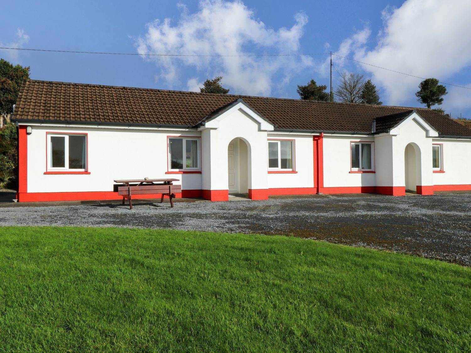 A white house with red trim and brown tiled roof with a wooden picnic table in front and green grass in the foreground at Robin's Roost 2 in Cornamona County Galway