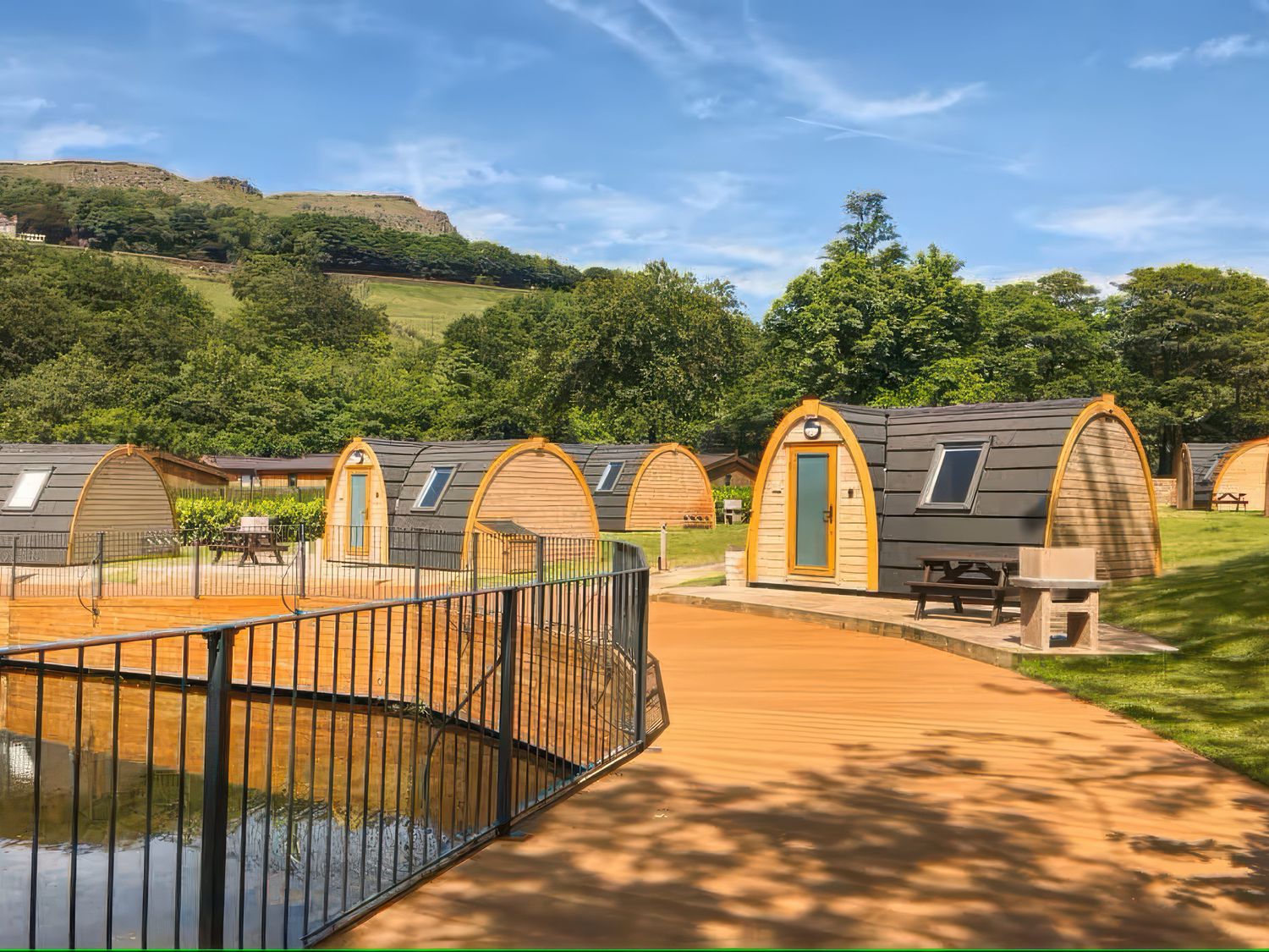 Small wooden pod cabins around a fenced pond with picnic tables on grass at Dovestone Pod 21 in Greenfield