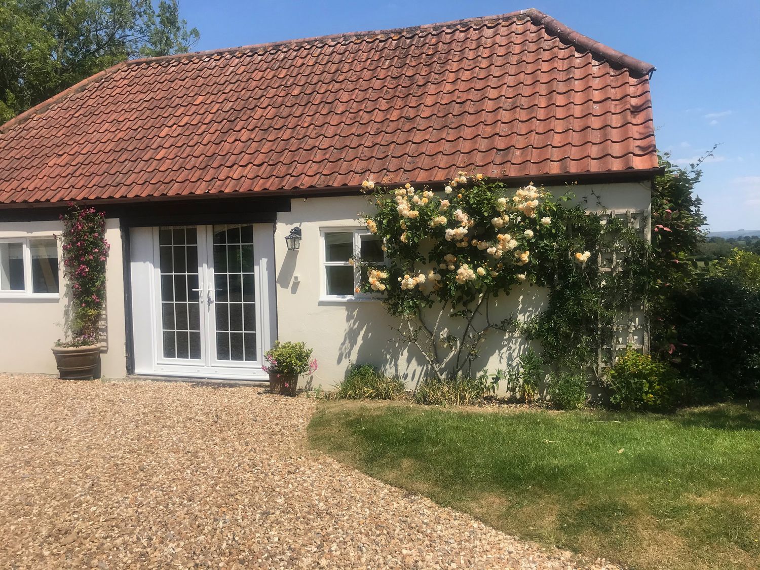 A small building with a red tiled roof white walls white double doors and climbing plants next to a gravel path and grass at Oke Apple Cottage in Okeford Fitzpaine