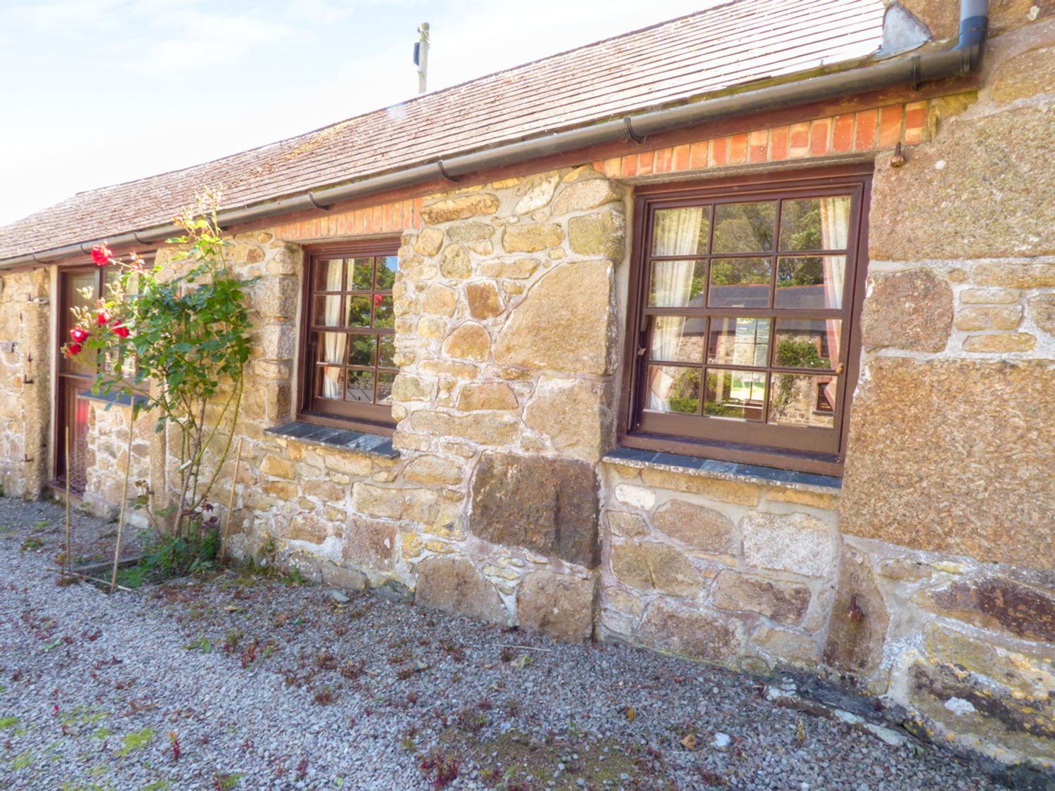 Exterior stone wall of a cottage with two brown framed windows and a door with a rose plant outside at Parlour Cottage in Marazion