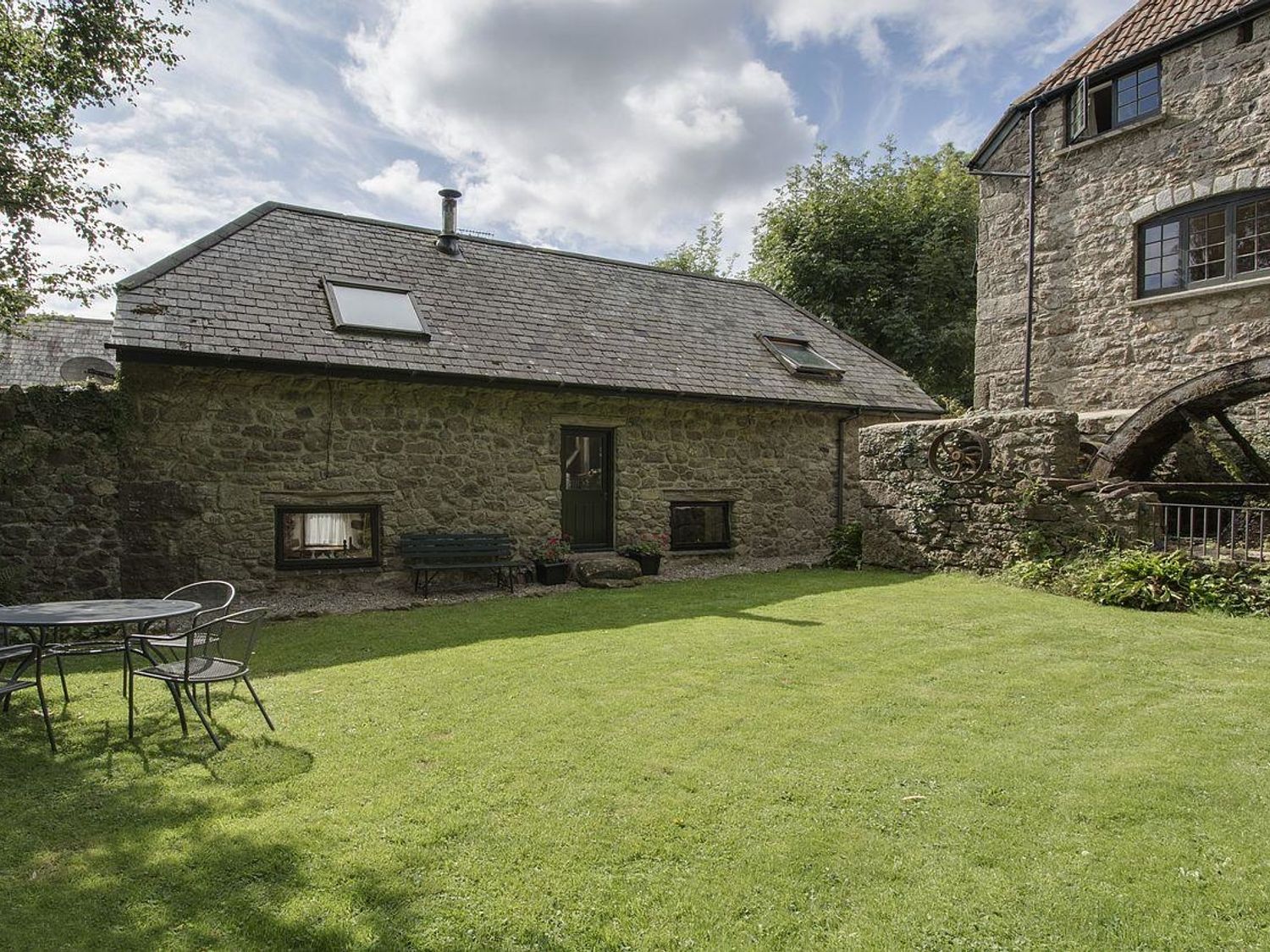 An outdoor lawn area with a round metal table and chairs next to a stone building with skylights and a water wheel at Water Barn Manaton near Lustleigh