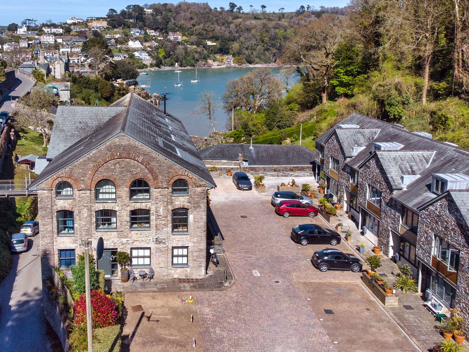 A paved parking area between two stone buildings with parked cars and a body of water surrounded by houses and trees in Dartmouth