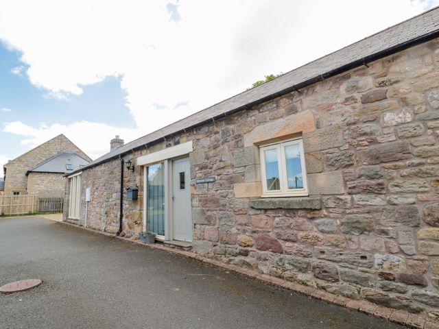 Exterior view of a stone cottage with a gray door and windows on a paved road at Hemmel Cottage in Chatton