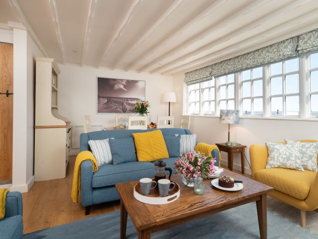 A living room with blue and yellow sofas a wooden coffee table with flowers and mugs and a dining table with chairs at Roxby Cottage in Staithes