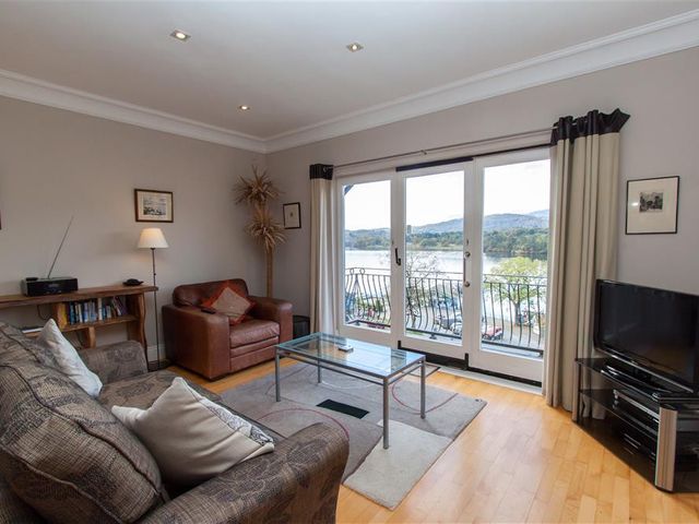 A living room with a sofa, armchair, glass coffee table, bookshelf, floor lamp, television, and large windows overlooking a lake at Lakeland View in Ambleside