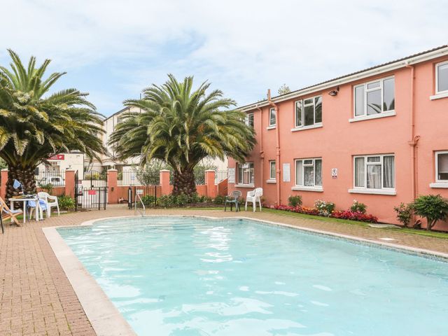 An outdoor swimming pool with chairs and palm trees next to a pink two-story building at Devon Seaside Apartment in Paignton