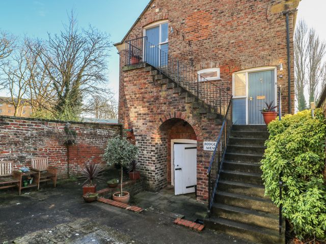 An outdoor area with brick building walls two sets of stairs potted plants wooden chairs and a door with a sign reading Rodgers Mews at 1 Rodgers Mews in Malton