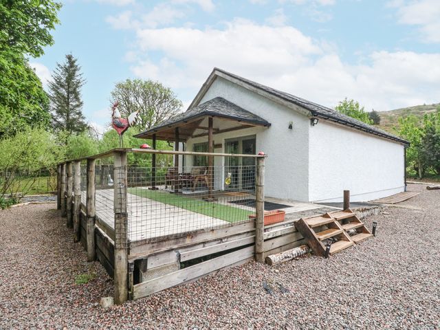 A white cottage with a wooden deck and metal railing surrounded by trees and gravel at Grant Cottage in Strontian
