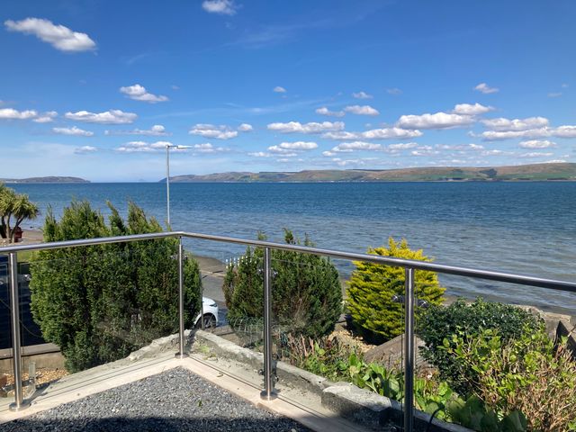 A balcony with glass railing overlooking the sea with hills in the distance and greenery below at Waters Edge in Stranraer