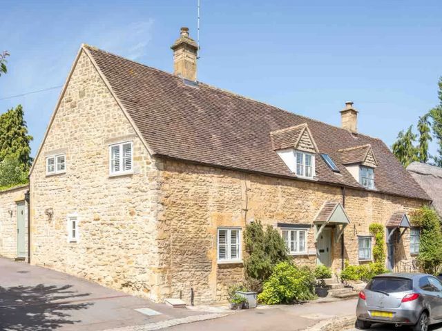 A stone cottage with a tiled roof and small windows with white shutters in a village setting at East Leaze in Chipping Campden