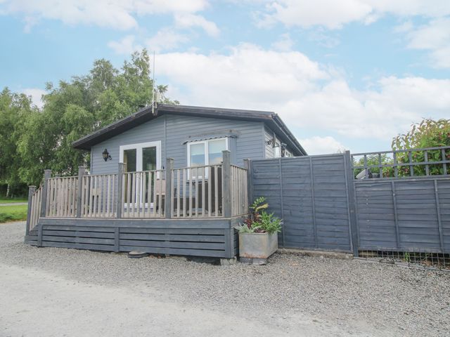 A gray wooden cabin with a fenced porch and plants outside at Wayside Lodge in Aston On Clun