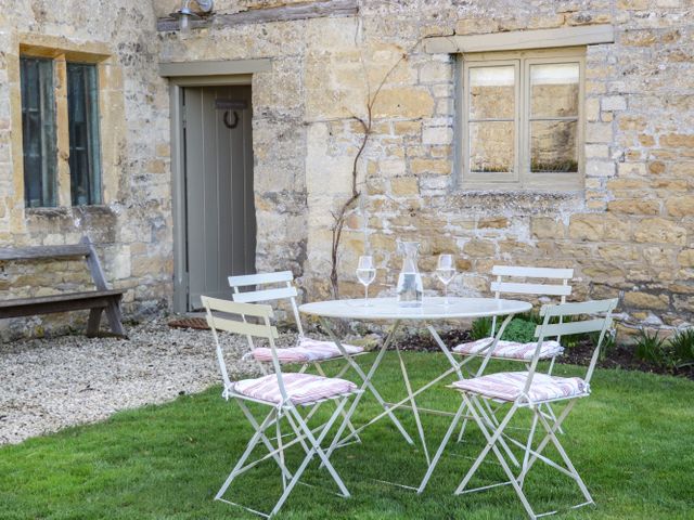 An outdoor area with a round table and four chairs with cushions on grass outside a stone building with windows and a door at The Compton Cottage in Northleach