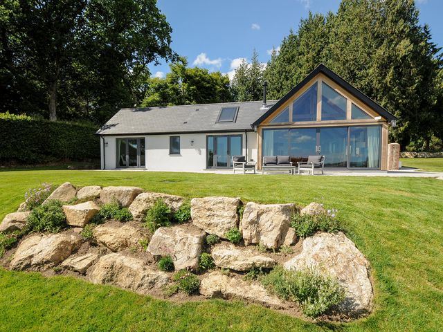 A modern house with large glass windows and a patio with outdoor chairs in a garden with rocks and grass at Sandsgate Lodge Spinster's Rock near Chagford