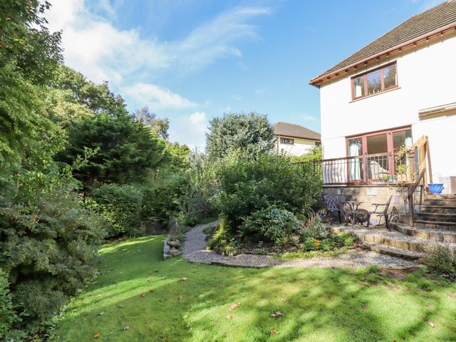 A garden with grass lawn shrubs and a stone pathway leading to a patio with chairs and a table at Taw River View in Sticklepath
