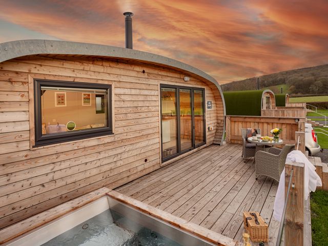A wooden cabin with a deck patio area featuring a hot tub a round table with chairs and a scenic view at Middle Longlands in Cartmel