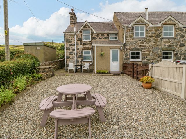 A gravel yard with a round wooden picnic table and two chairs outside a stone cottage at Moonlight Cottage in Sarn Meyllteyrn