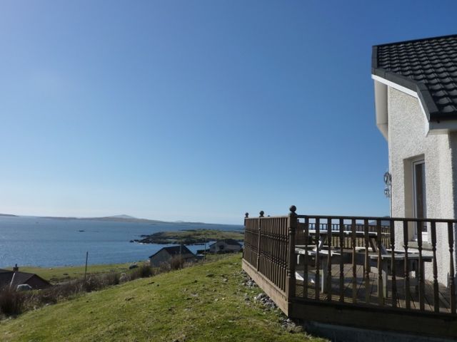A house with a wooden deck overlooking a body of water and grassy land at Shalom Cottage in Leverburgh