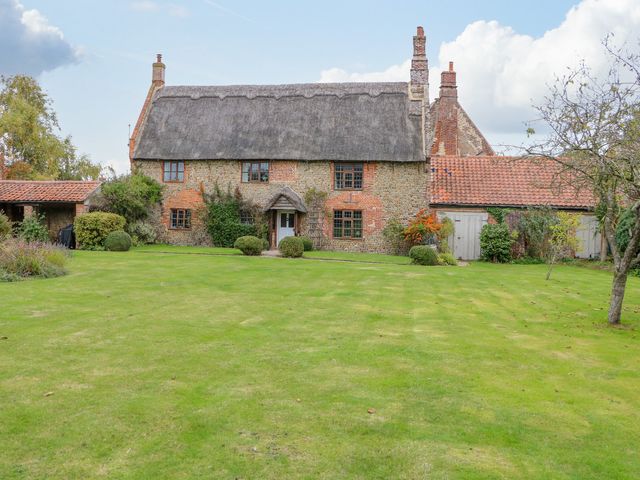 A stone and brick house with a thatched roof and chimneys surrounded by a lawn and shrubs at Manor Farm House in Bacton