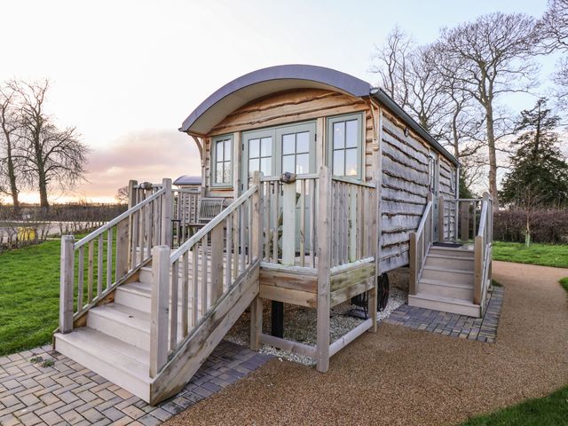 A wooden shepherd hut with two staircases and a small porch surrounded by trees and grass at The Storyteller in Bolton Percy near Tadcaster