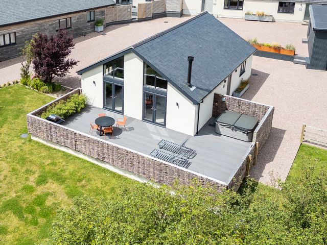 A small house with a fenced patio area containing a round table with orange chairs two lounge chairs and a hot tub surrounded by grass and other buildings at Garden House in Llanfechell