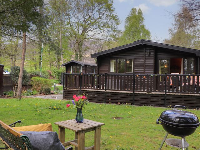 A garden with a wooden bench and table with flowers and a charcoal grill near a dark wooden cabin at Derwent Lodge Keswick