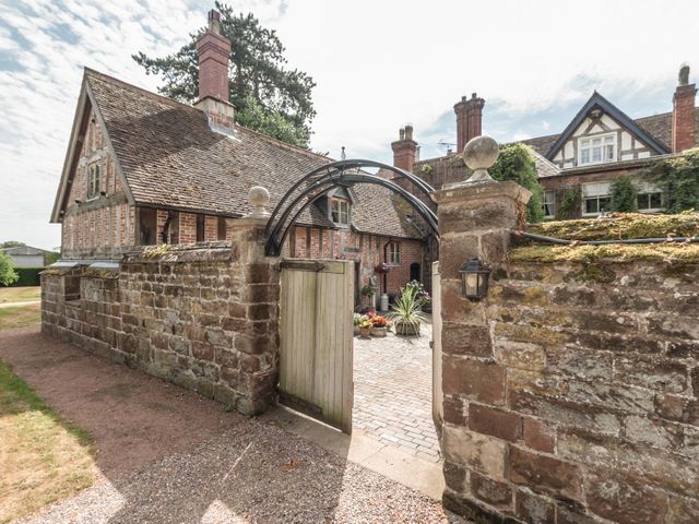 A stone wall with an open wooden gate leading to a courtyard with potted plants and brick buildings