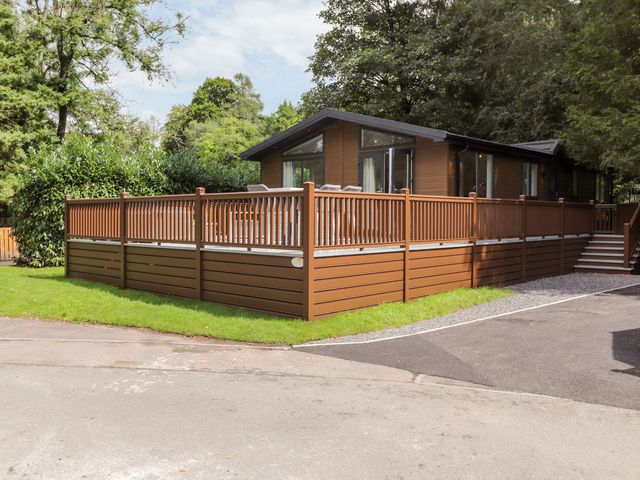 A brown wooden lodge with a fenced deck surrounded by trees and greenery at Unique Luxury Lodge 1 Langdale White Cross Bay near Windermere