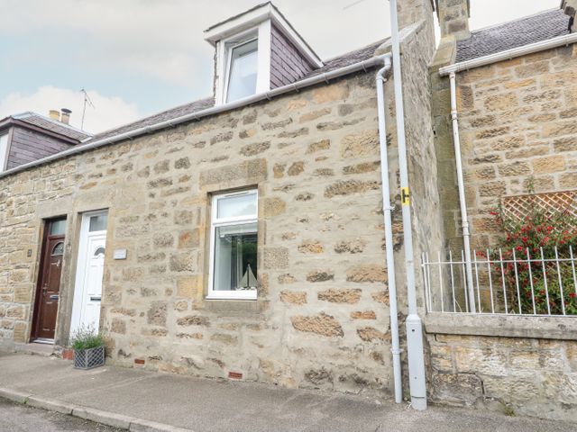 Stone exterior of a cottage with white and brown doors and a window with a model sailboat at Anchor Cottage in Hopeman