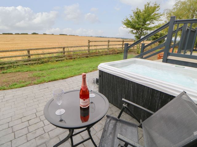 An outdoor patio with a table holding a bottle and two glasses next to a hot tub and a chair overlooking a fenced field at Bowes Hideaway in Eppleby