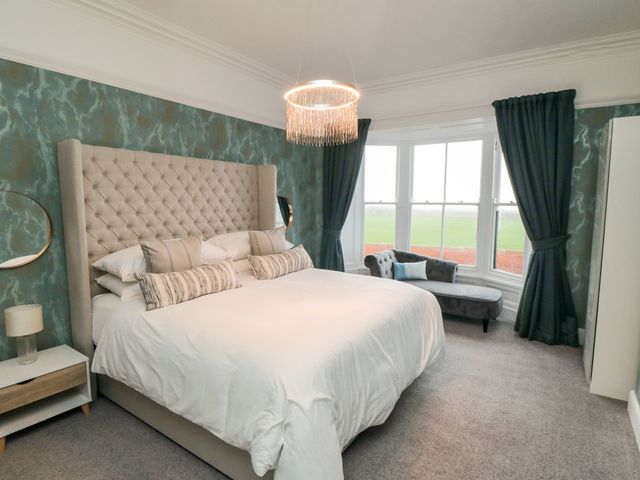 A bedroom with a large bed white bedding tufted headboard two pillows a side table lamp a chaise lounge under bay window with dark curtains at No.20 The Headland in Hartlepool