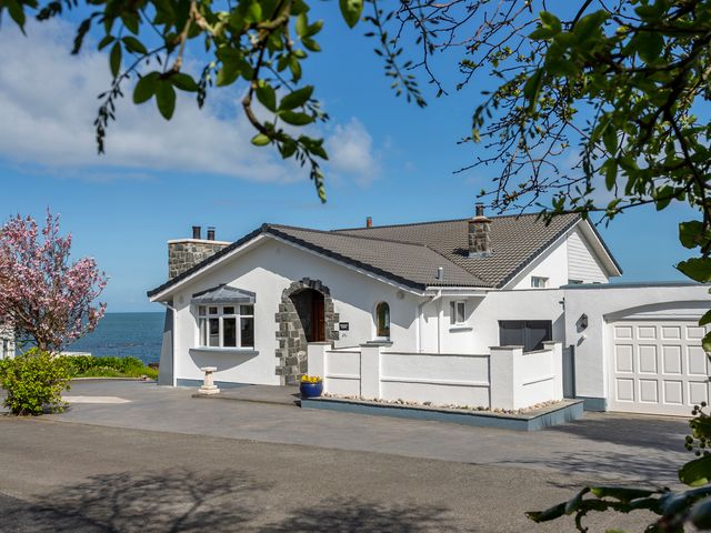 A white house with a stone arch entrance and a garage near the sea with trees and a pink flowering bush at Rocklyn Lodge in Donaghadee