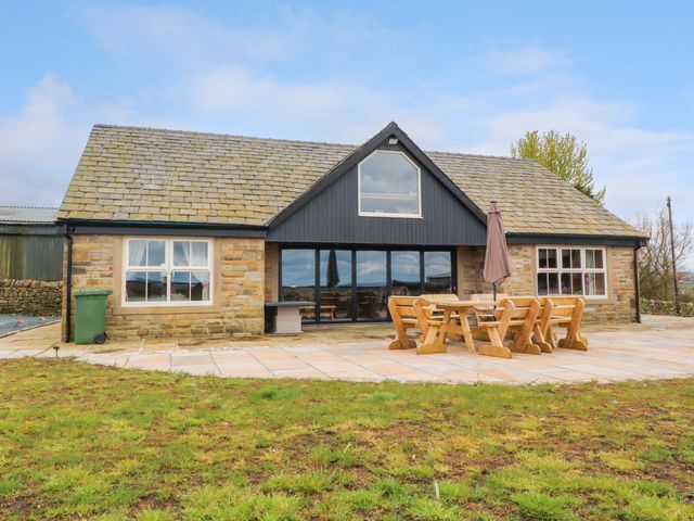 A stone house with a tiled roof and large glass doors with wooden patio furniture on a paved area in front of it at Meadow Top Farm Barn in Clitheroe