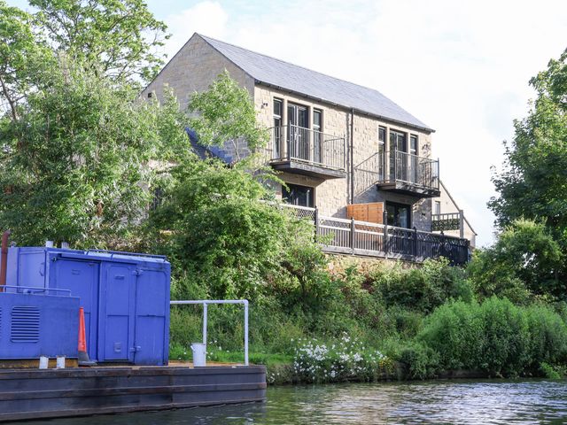 A stone house with balconies and black railings near a river with green bushes and a blue structure on the riverbank at Waters Edge Lock View in Gargrave