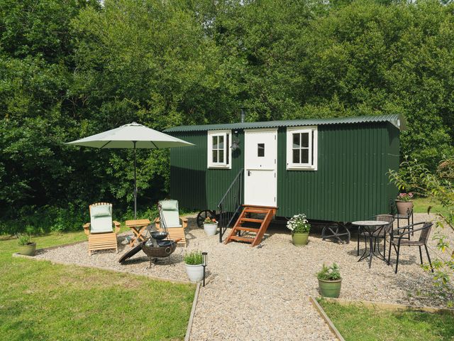 A green shepherd hut with white door and windows on a gravel area with chairs tables and an umbrella at Collen in Aberaeron