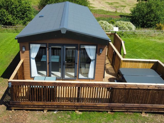 A small wooden cabin with glass doors on a wooden deck with patio furniture and a covered hot tub in a grassy area at Willow Lodge in Whitby